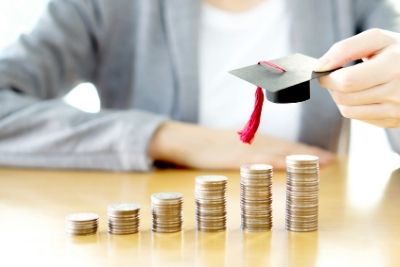 A row of stacked coins, increasing in size, on a table. A person is in the background is holding a miniature graduation cap over the largest pile of coins.