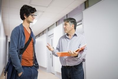 A tall male student with glasses, speaking with a male professor while in a bright hallway.