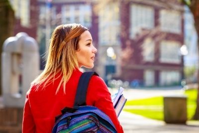 A woman approaching a post-secondary building, looking over her right shoulder with a contemplative expression.