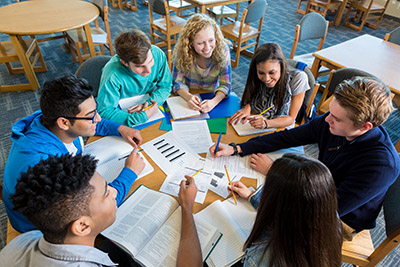 7 students sitting at a round table, discussing while working with a variety of papers scattered at their table.