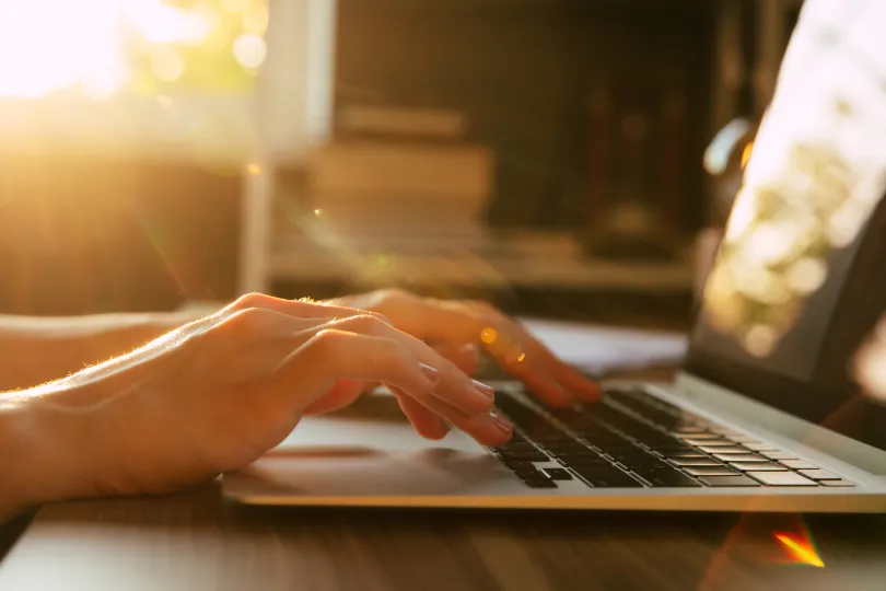 A close-up shot of hands typing on a laptop keyboard, bathed in warm sunlight