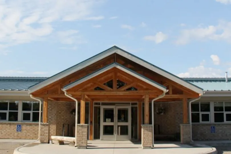 An exterior view of the Kenjgewin Teg entrance, showcasing a light-colored brick building with a large wood-beamed portico and a peaked roof