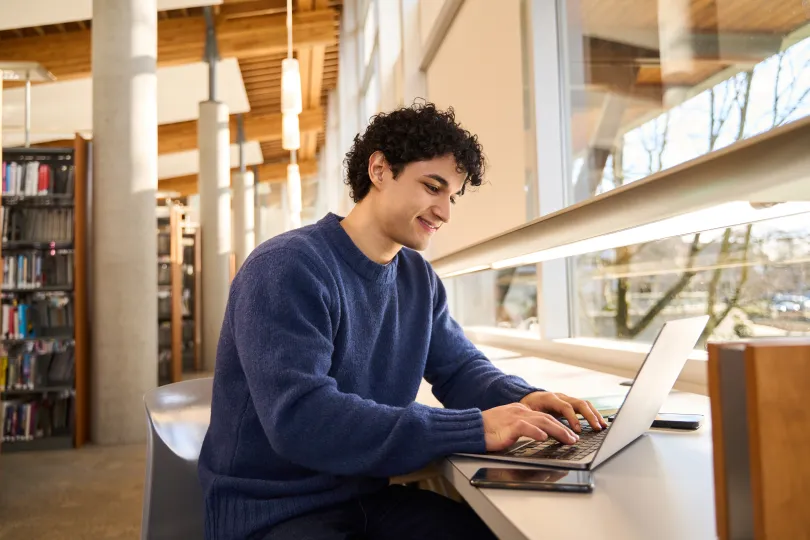 University student happily studying with a laptop in a bright library, surrounded by books and natural light
