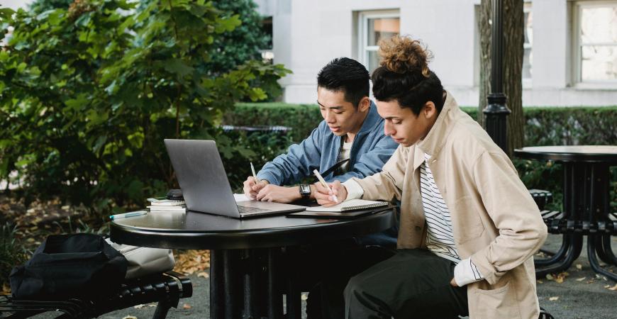 Two students sit at an outdoor table, taking notes, with a laptop open in front of them.