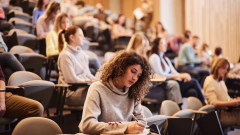 Female college student writing an exam during a class at lecture hall. Her classmate are in the background.