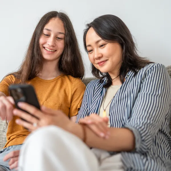 Mother and teenage daughter relaxing on a sofa, sharing a moment while using a smartphone together.