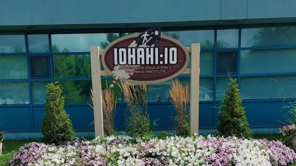An outdoor sign for the Iohahi:io Akwesasne Education & Training Institute, featuring a large feather graphic, situated in front of a blue building and a garden of purple and white flowers